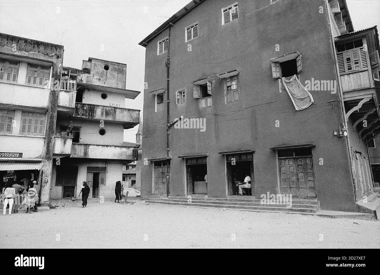 Zanzibar, Tanzania - Una fotografia in bianco e nero raffigura una scena di strada a Stone Town, Zanzibar. Numerosi edifici, costruiti in mattoni rossi e caratterizzati da dettagli architettonici tradizionali, fiancheggiano la strada. Le persone sono visibili davanti agli edifici, suggerendo un ambiente urbano attivo. La fotografia evidenzia il carattere storico e architettonico della città. Foto Stock