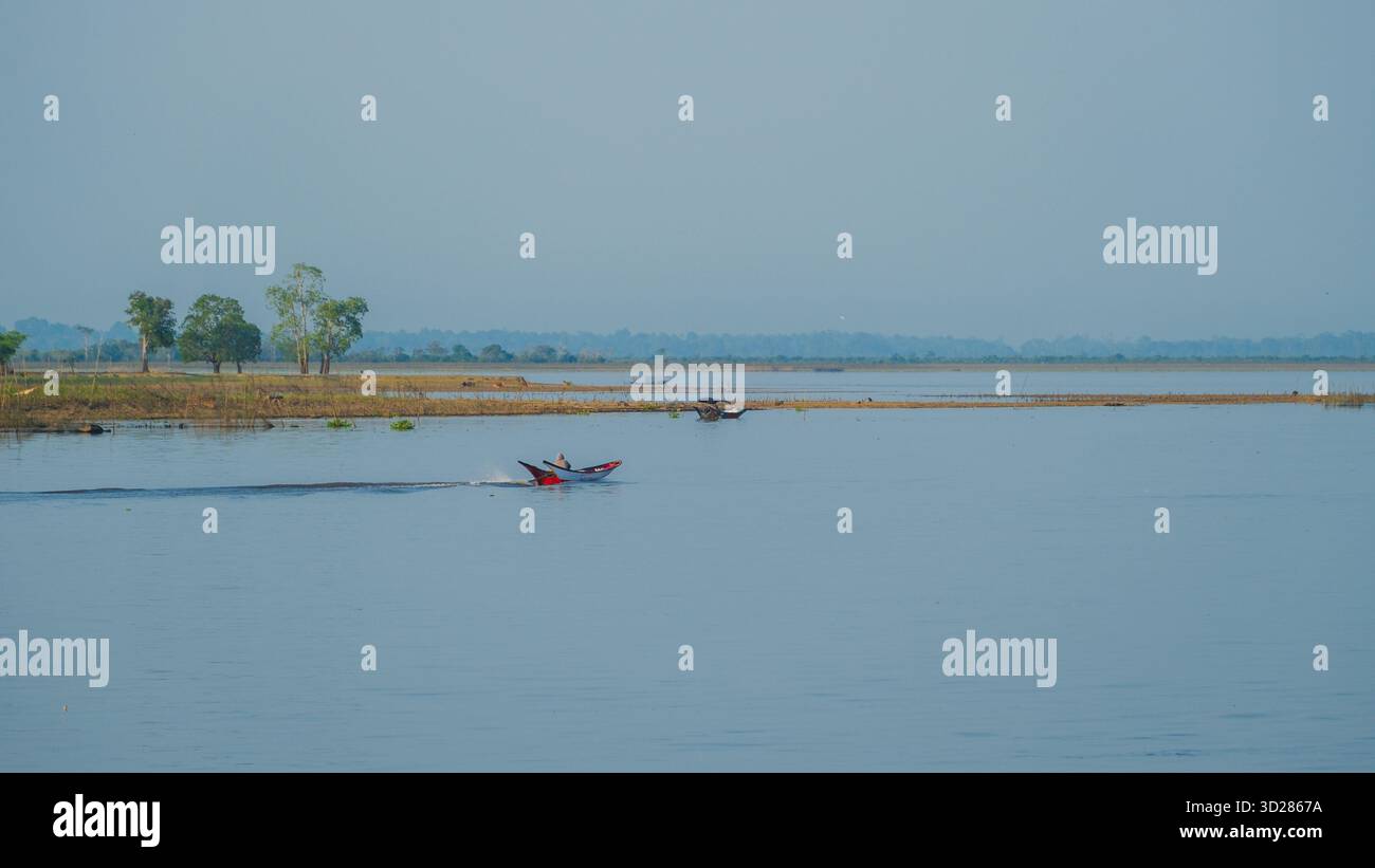 Un pescatore guida un piccolo motoscafo attraverso un fiume tranquillo circondato da paludi naturali e alberi lontani. Tranquillo paesaggio rurale che mostra la tradizione Foto Stock