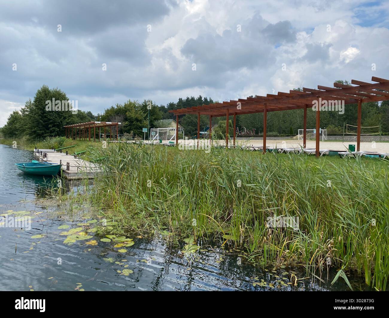 Una tranquilla scena sul lago in una giornata nuvolosa, con una piccola barca verde legata ad un molo di legno. Sullo sfondo, una spiaggia sabbiosa con lettini Foto Stock
