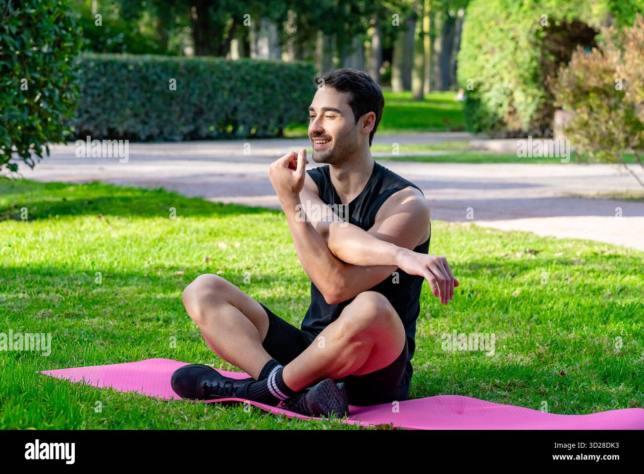 Uomo che esegue uno stretching del braccio seduto a gambe incrociate su un tappetino da yoga in un parco Foto Stock