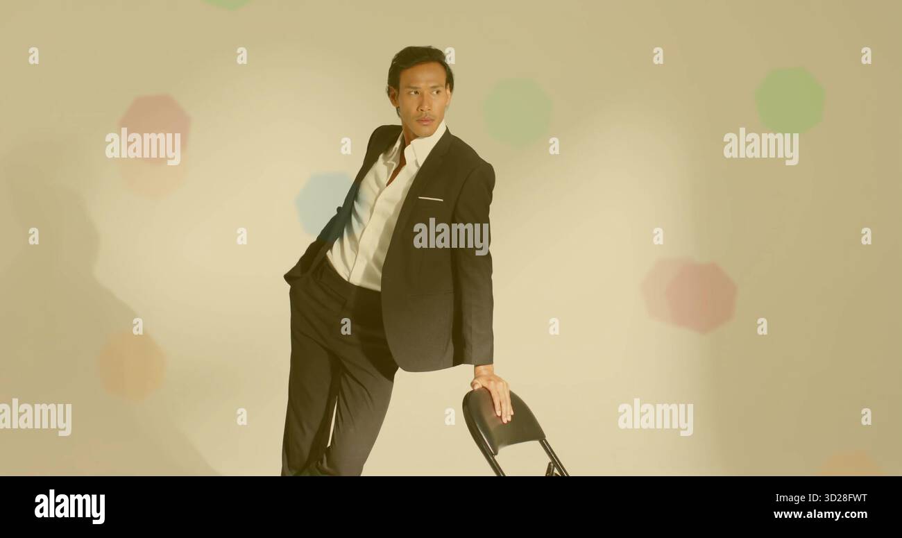 Modello pendente che indossa una tuta nera e una camicia bianca che guarda a sinistra in studio pastello, con sedia pieghevole Foto Stock