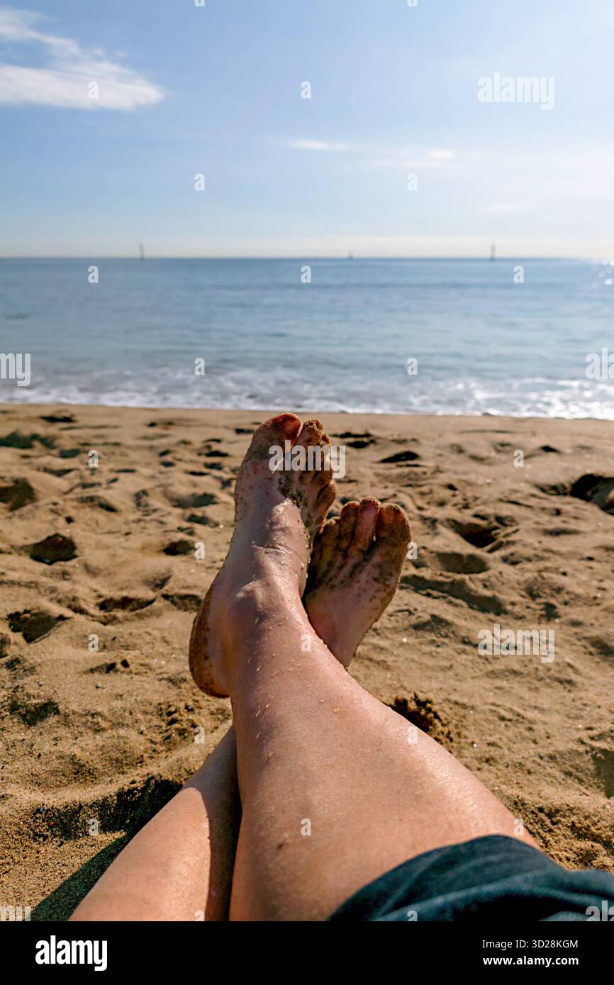 I piedi di una persona sono nella sabbia della spiaggia. La persona è sdraiata sulla spiaggia e l'acqua è calma. La spiaggia e' sabbiosa e il mare e' nella parte posteriore Foto Stock