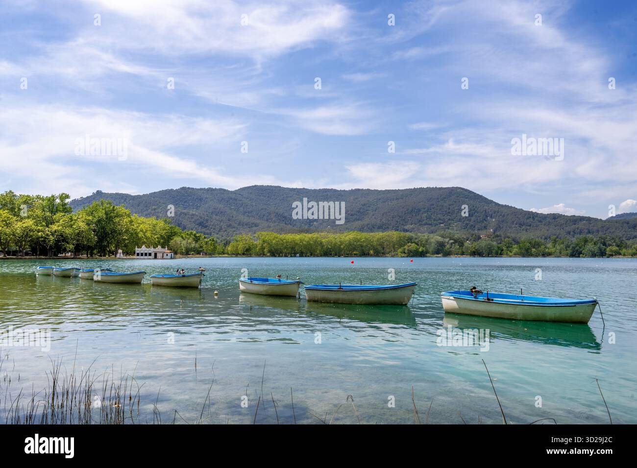 Barche a remi che galleggiano sulle calme acque turchesi del lago Banyoles, Catalogna, Spagna, con montagne sotto un cielo blu Foto Stock