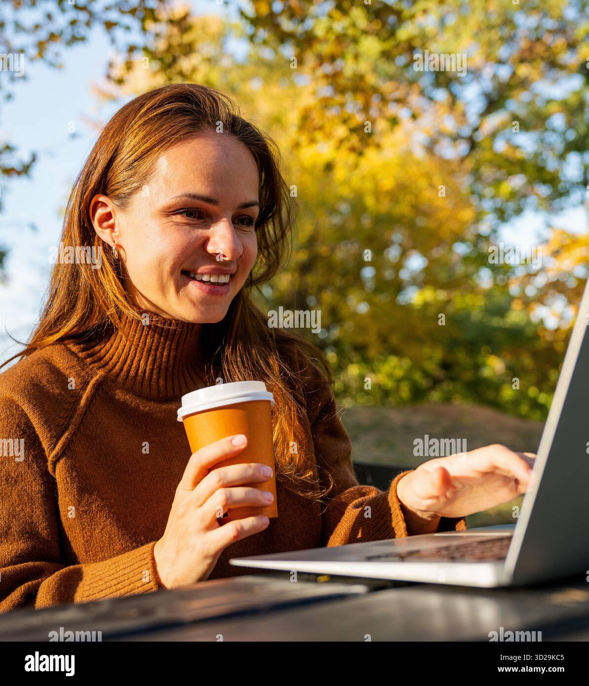 Donna sorridente con caffè da asporto che lavora su un notebook all'aperto nel parco autunnale. Foto Stock