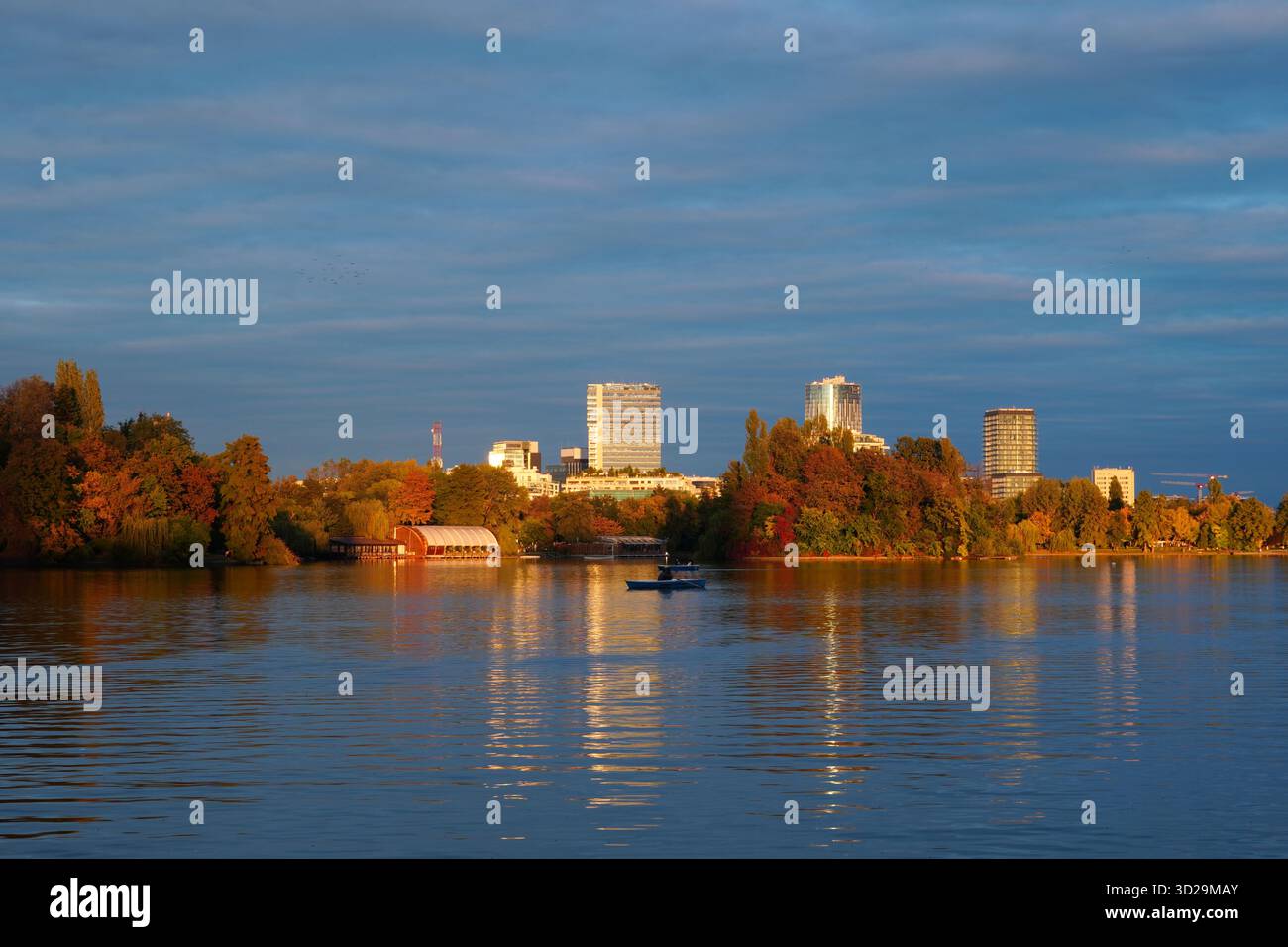 Splendido paesaggio autunnale all'ora d'oro nel parco Herastrau di Bucarest con barche sul lago, alberi colorati e torri di uffici in lontananza Foto Stock