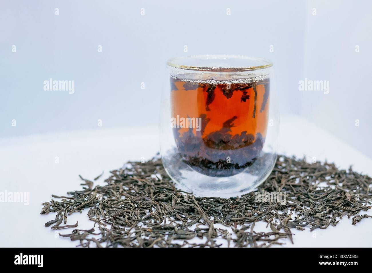 Tazza di vetro con tè nero estratto, circondata da foglie di tè sciolte su una superficie bianca. Pubblicità di bevande, promozione benessere, pacchetto di prodotti biologici Foto Stock