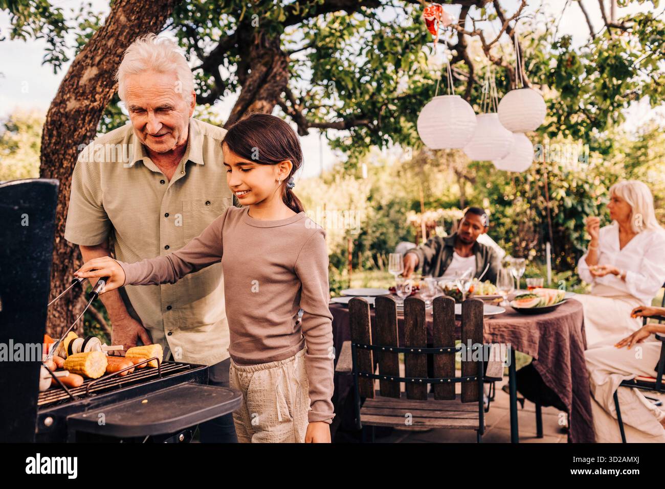 Ragazza sorridente che grigia il cibo al barbecue con l'aiuto del nonno alla festa in giardino Foto Stock