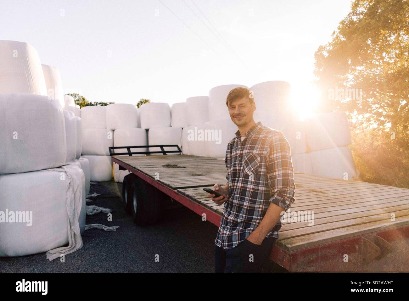 Ritratto di un agricoltore sorridente che si appoggia a un rimorchio con smartphone Foto Stock