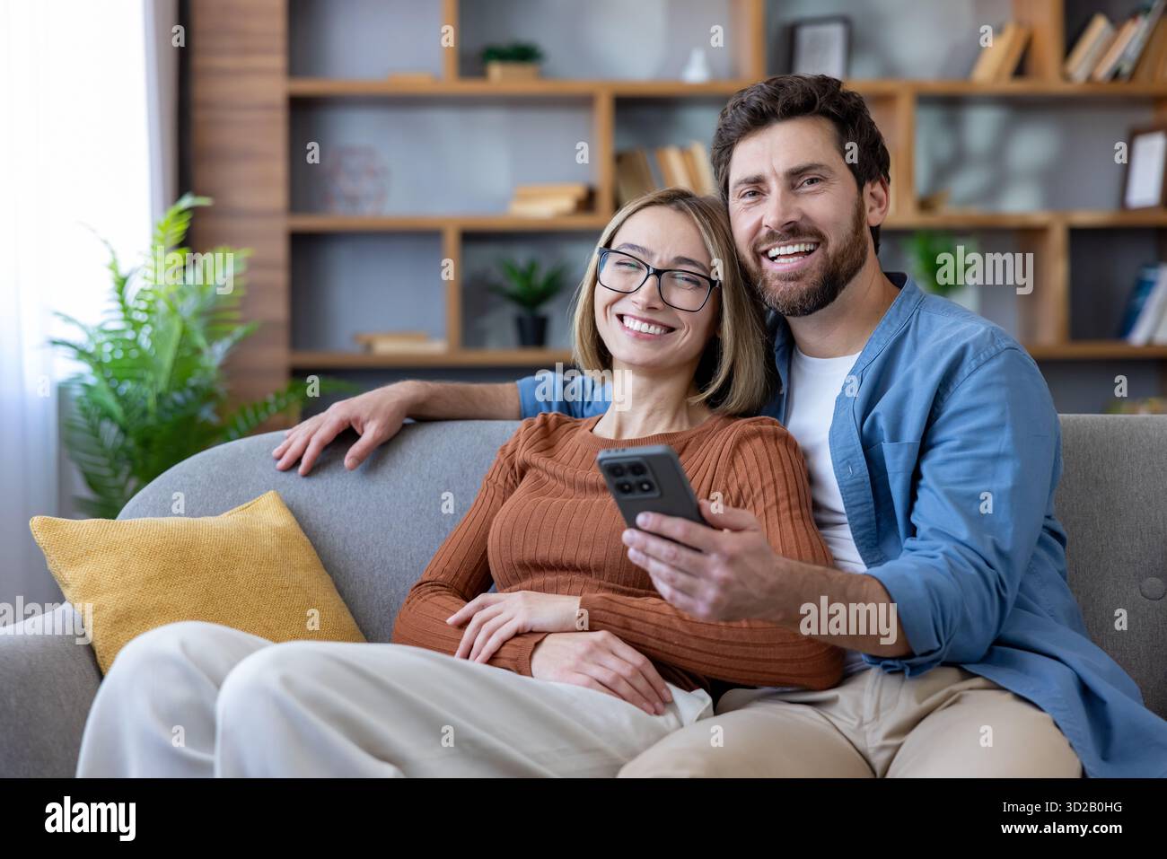 Coppia seduta su un comodo divano, ridendo e sorridendo mentre posa per un autoritratto utilizzando un telefono cellulare, godendosi un momento informale a casa Foto Stock