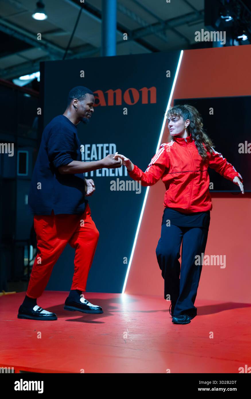 12.10.2025 Salon de la Photo Paris.Danza scintillante: Uomo e donna in abiti rossi/neri, tenendo per mano un palco rosso con il logo Canon. Foto Stock
