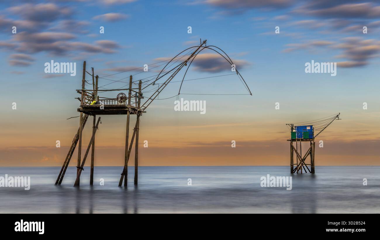 Due piattaforme di pesca in legno a Plage des Dunes, Francia, si stagliano sulle acque calme durante un tranquillo tramonto. Il cielo è adornato da morbide nuvole e sfumature Foto Stock