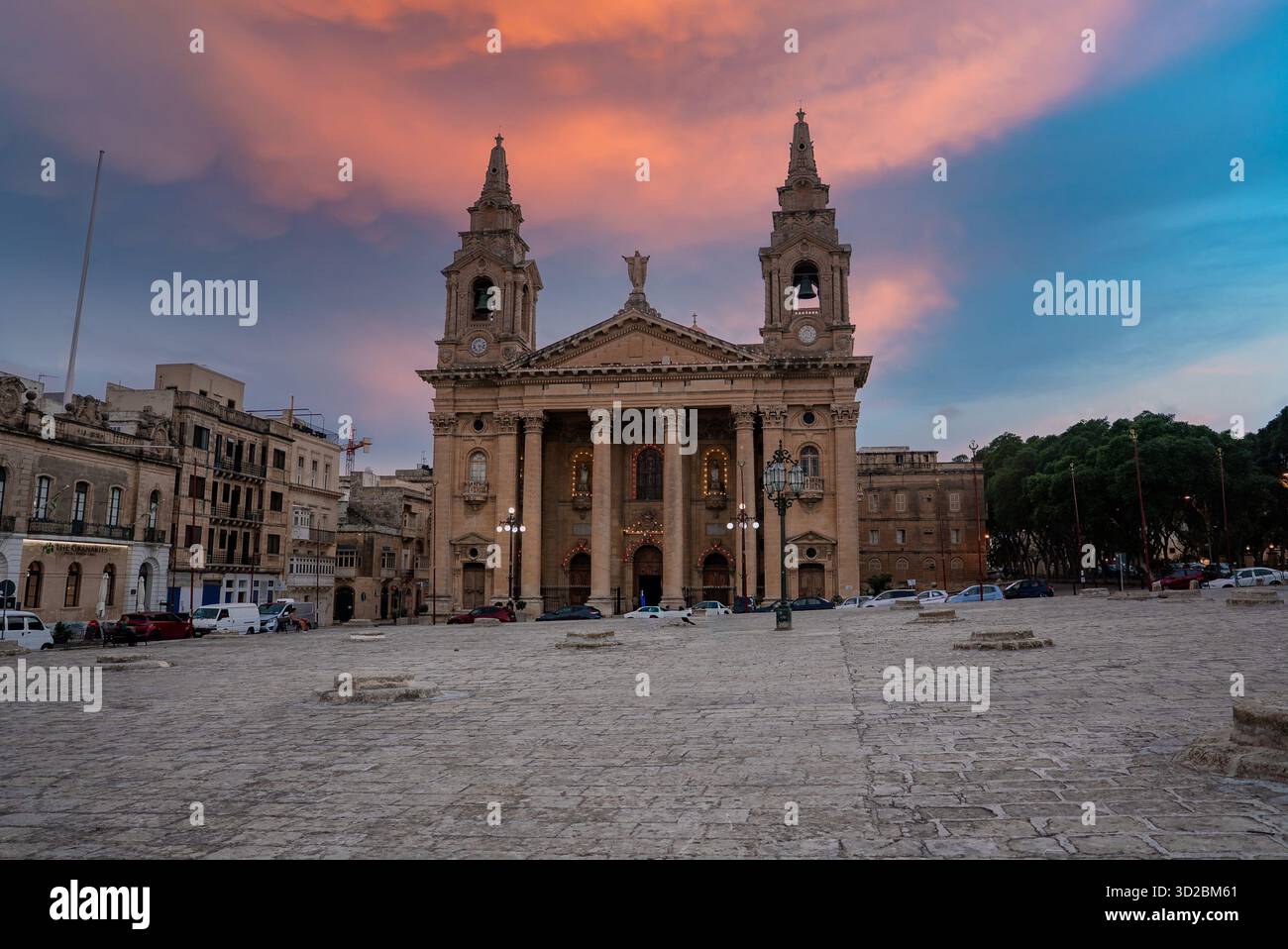 Una chiesa barocca con torri gemelle e un portico si affaccia su una piazza in pietra a la Valletta, Malta al tramonto. Una luce calda si illumina sulla pietra calcarea, parcheggio auto nelle vicinanze, la Foto Stock