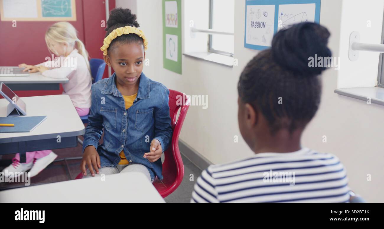 Ragazza seduta con fascia gialla e giacca in denim alla scrivania della scuola, tablet e poster Foto Stock