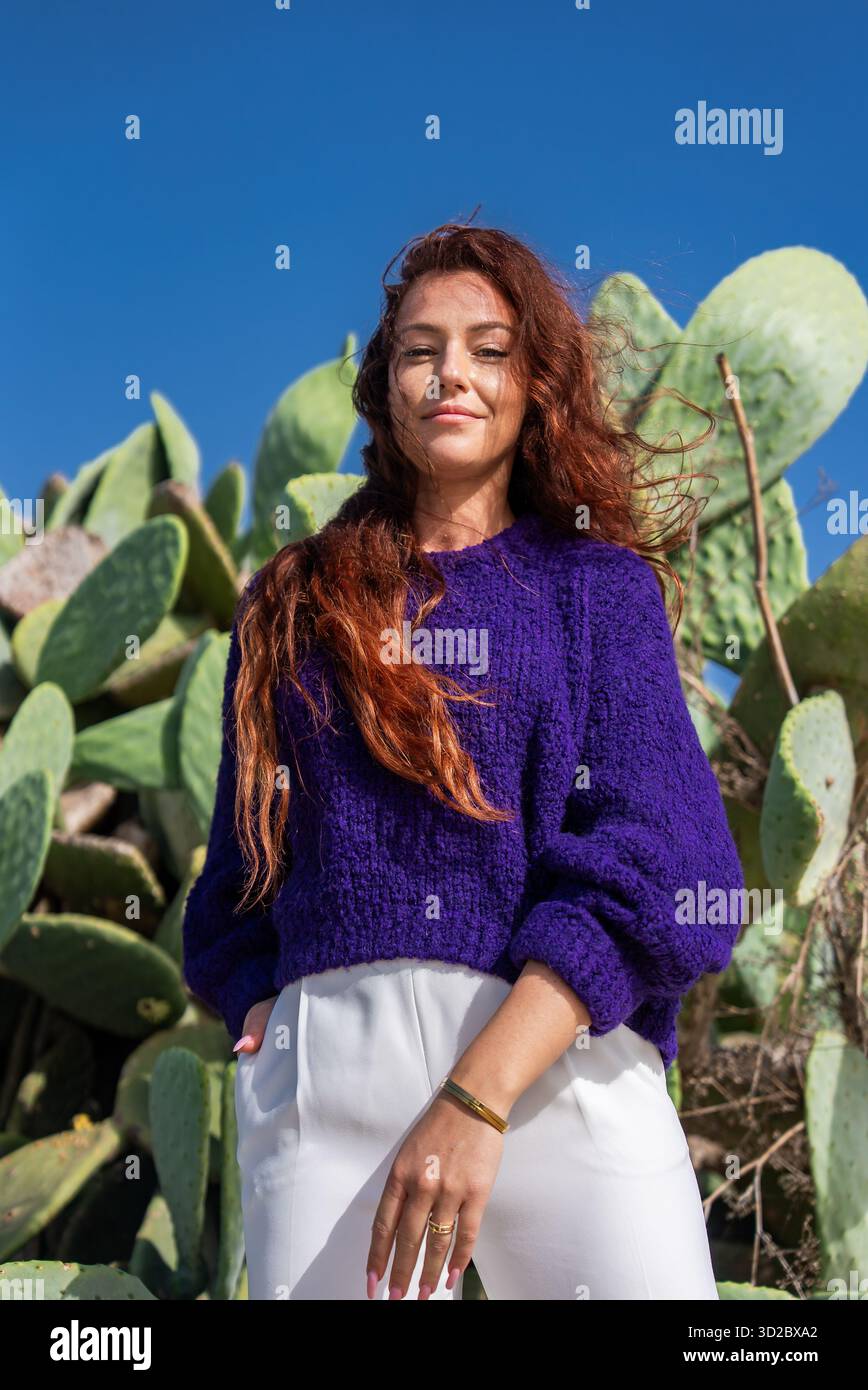 Una donna con i capelli di auburn sta davanti ai cactus di fichi d'India a Mellieha, Malta, mani in tasca, luce di mezzogiorno, angolo basso verso l'alto, viola e verde audaci Foto Stock