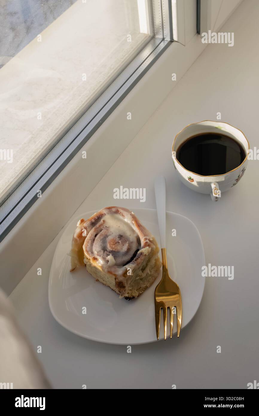 Un ambiente accogliente caratterizzato da un rotolo di cannella appena sfornato su un piatto bianco con forchetta d'oro, accanto a una tazza di caffè nero su un davanzale. Foto Stock