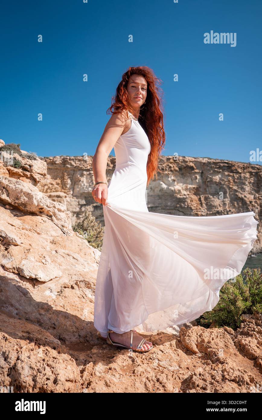 Una donna solleva il suo vestito bianco sulle scogliere calcaree vicino alla Laguna Blu a Comino, Malta. Luce del giorno intensa, colori vivaci, angolo basso verso l'alto, movimento del tessuto Foto Stock