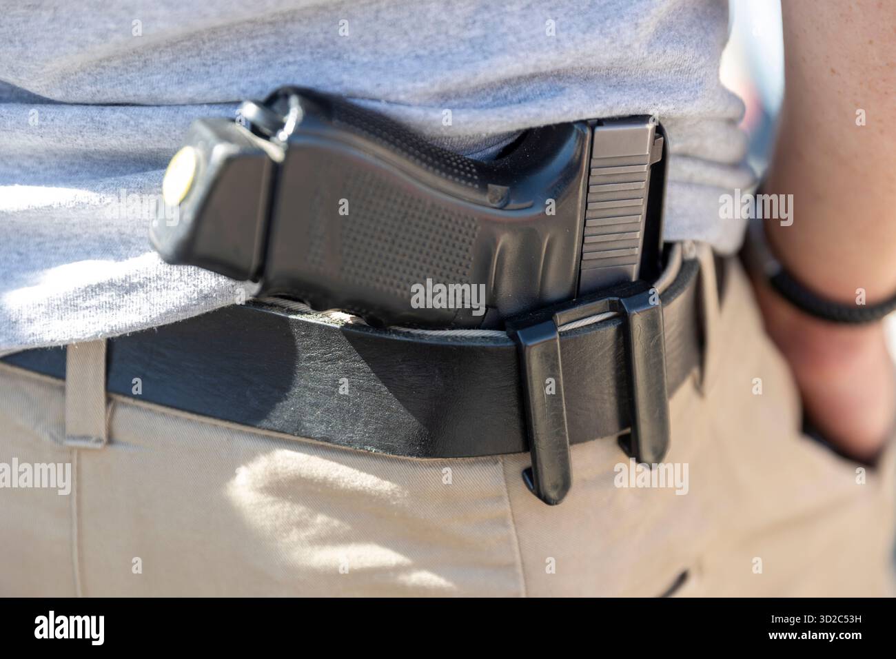 Uno scatto ravvicinato e discreto di una pistola nera infilata in una custodia indossata su una cintura nera da una persona che indossa pantaloni tan e una camicia grigia. Questo suggerisce Foto Stock