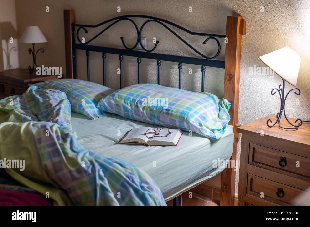 Un'accogliente camera da letto presenta una struttura del letto in metallo, biancheria da letto a quadri, un libro aperto con bicchieri da lettura e calde lampade da comodino che creano una tranquilla e familiare atmos Foto Stock