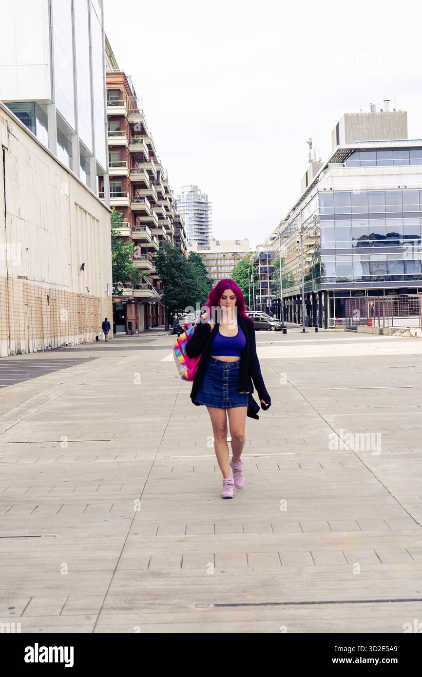 Giovane donna con i capelli viola che cammina con sicurezza in un paesaggio urbano moderno, portando con sé uno zaino colorato. Concetto di moda urbana Foto Stock