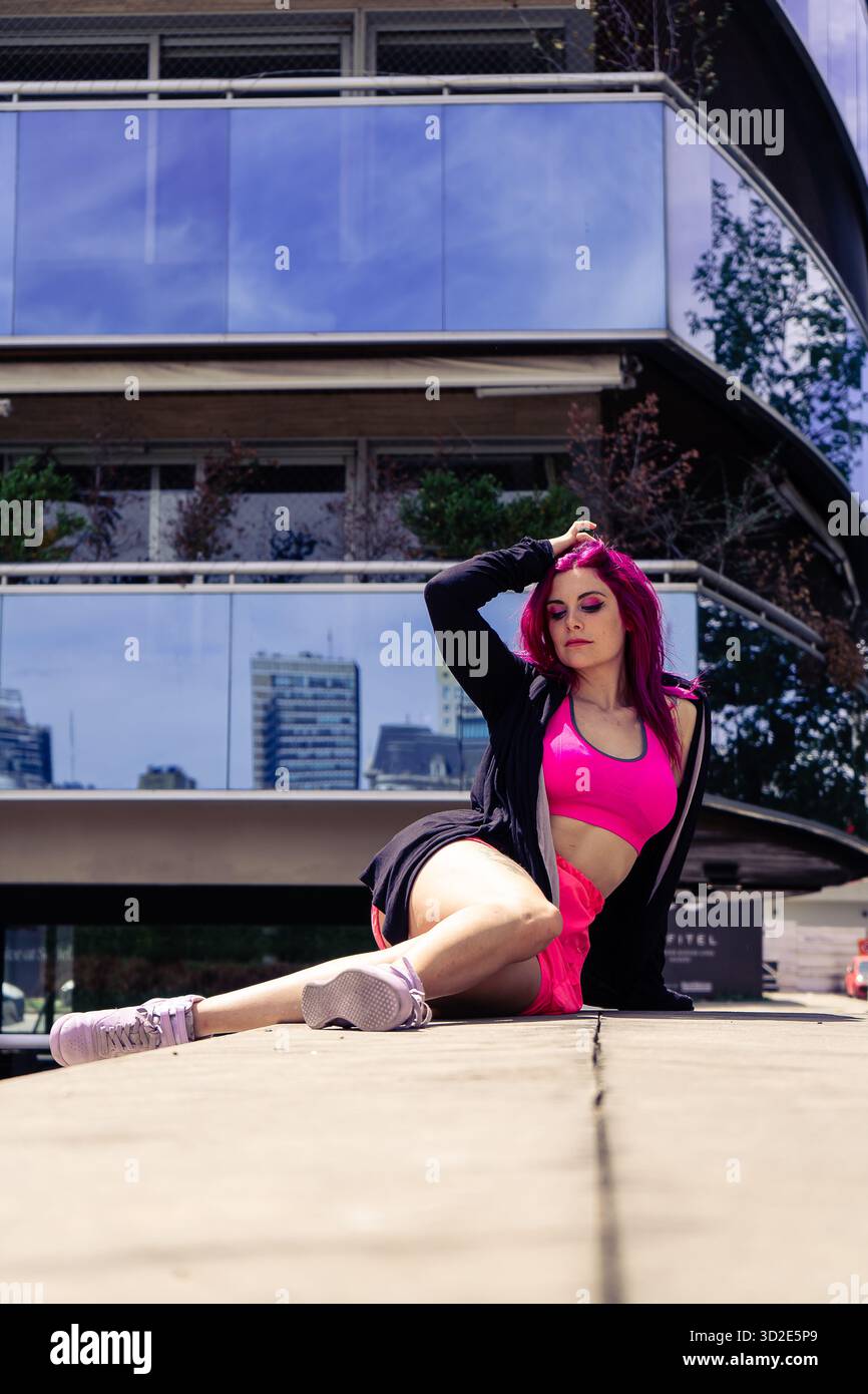 Donna dai capelli rosa che indossa un abbigliamento sportivo in posa in un ambiente urbano con architettura e riflessi moderni Foto Stock