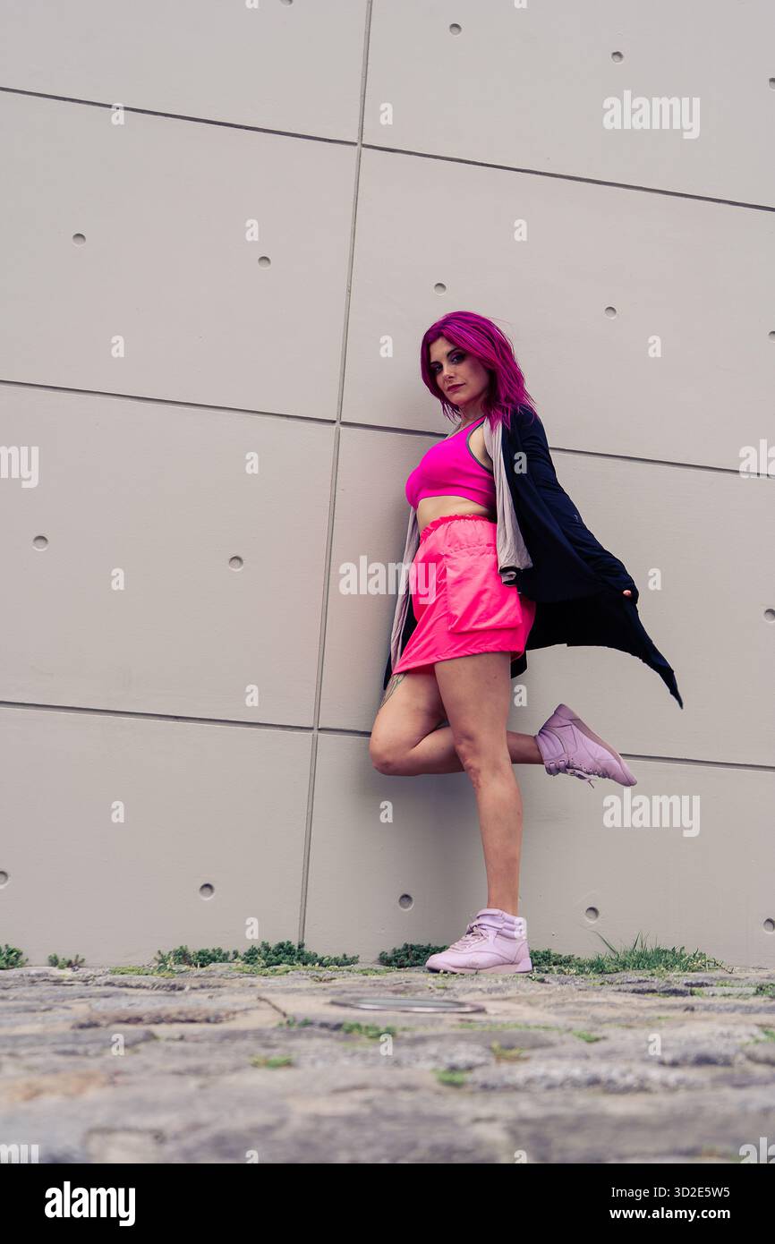 Giovane donna sicura di sé con vivaci capelli rosa e abbigliamento sportivo luminoso che si posa contro una moderna parete urbana in cemento Foto Stock