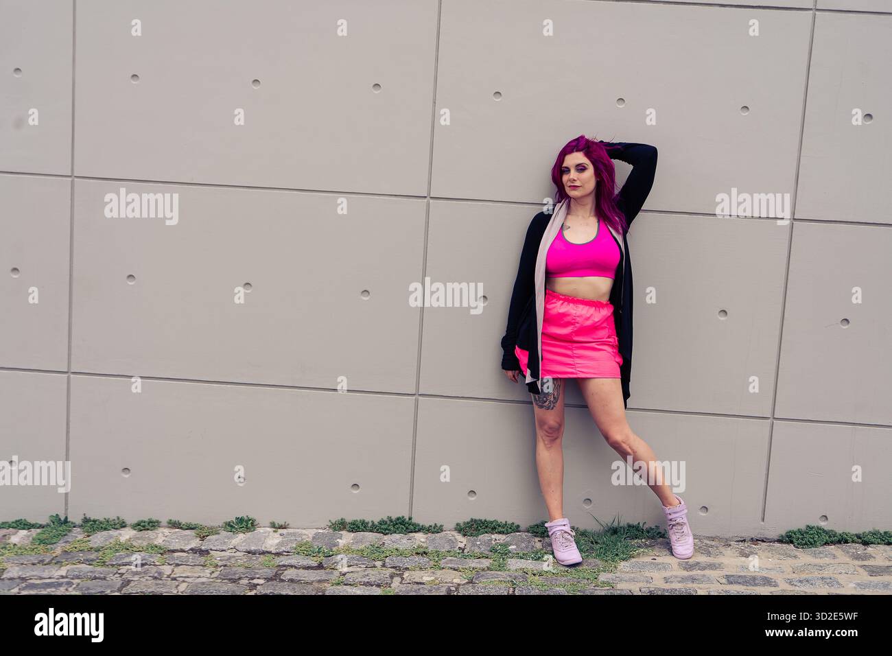 Giovane donna che sfoggia una moderna Street fashion con capelli e outfit rosa vivaci, appoggiata con sicurezza a un muro urbano minimalista Foto Stock