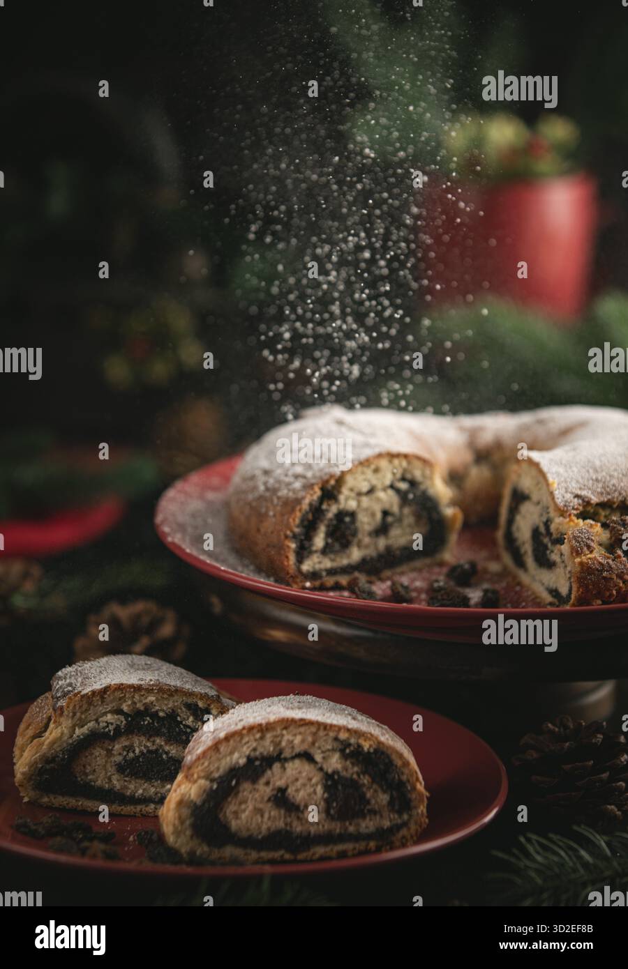 Lo zucchero a velo cade sulla torta di semi di papavero su un piatto rosso circondato da decorazioni natalizie Foto Stock