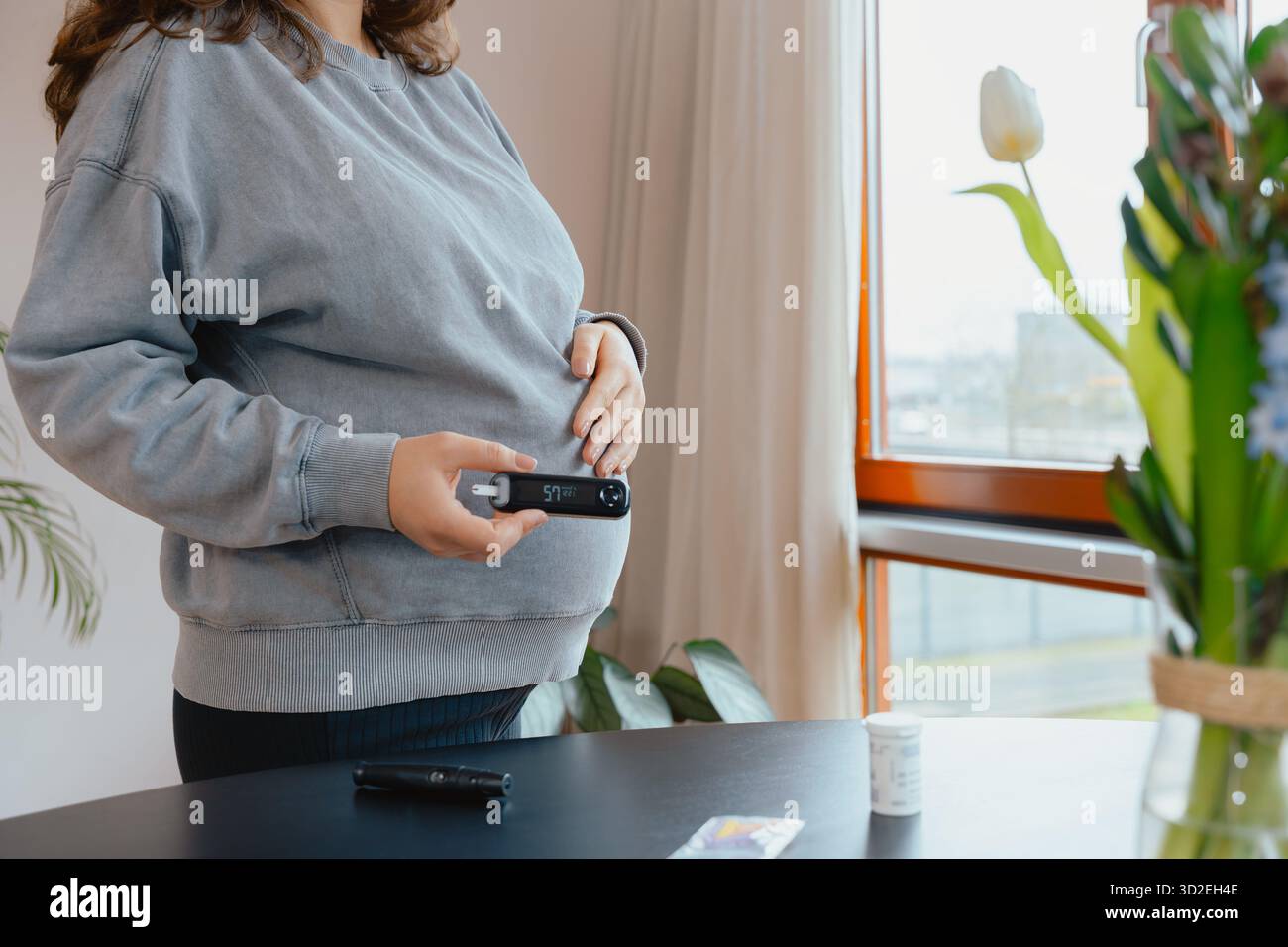 Una donna incinta sta accanto alla finestra, tenendo un glucometro e toccandole la pancia mentre monitora la glicemia per la cura del diabete gestazionale Foto Stock