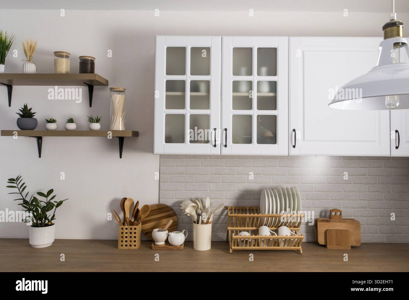 Parte dell'interno di una moderna cucina bianca con armadi in vetro smerigliato, una parete bianca in mattoni e un ripiano in legno. attrezzature da cucina Foto Stock