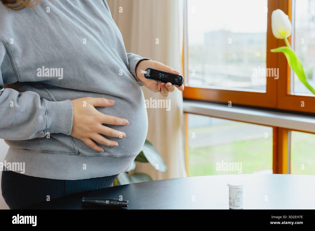 Una donna incinta tiene un glucometro mentre riposa l'altra mano sulla pancia, monitorando i livelli di zucchero nel sangue durante la cura del diabete gestazionale Foto Stock