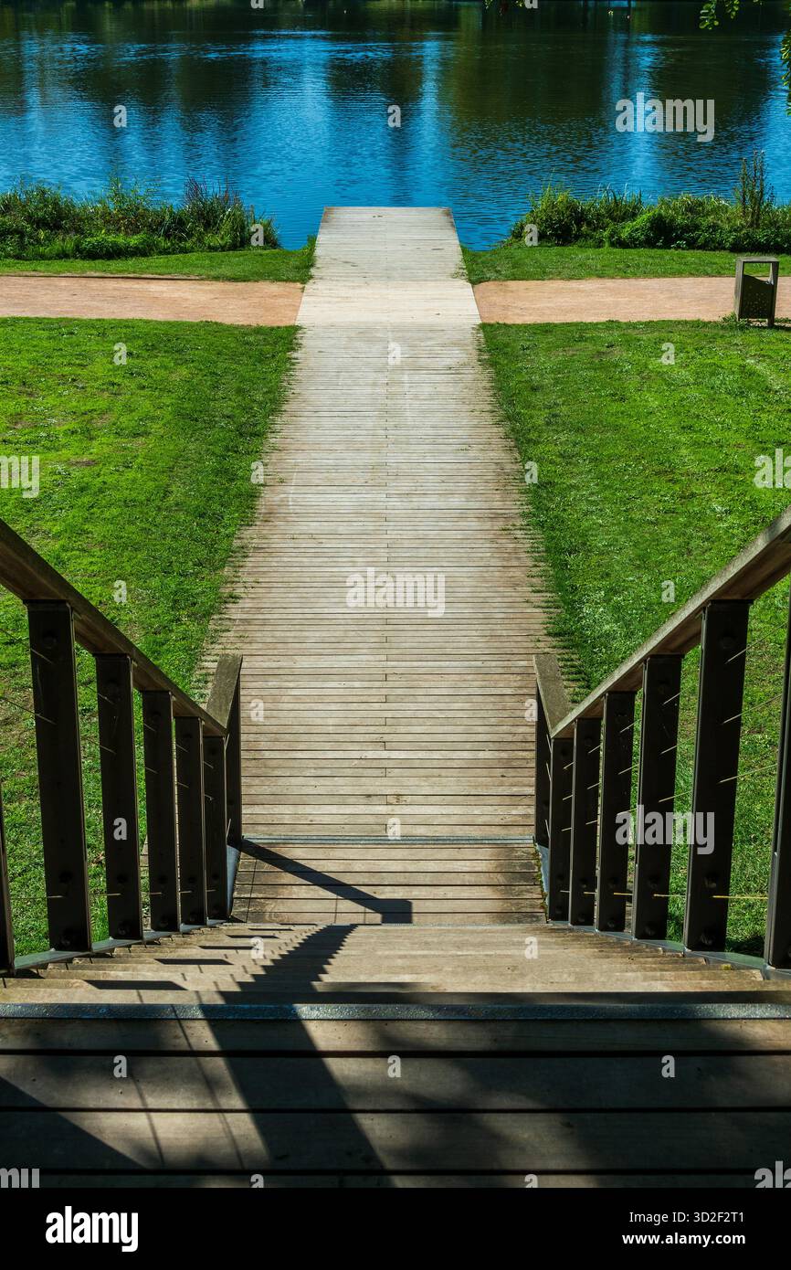 Le scale conducono a un sentiero di legno attraverso l'erba verde verso un lago calmo. Un molo si estende nell'acqua. Vichy. Allier. Auvergne. Francia Foto Stock