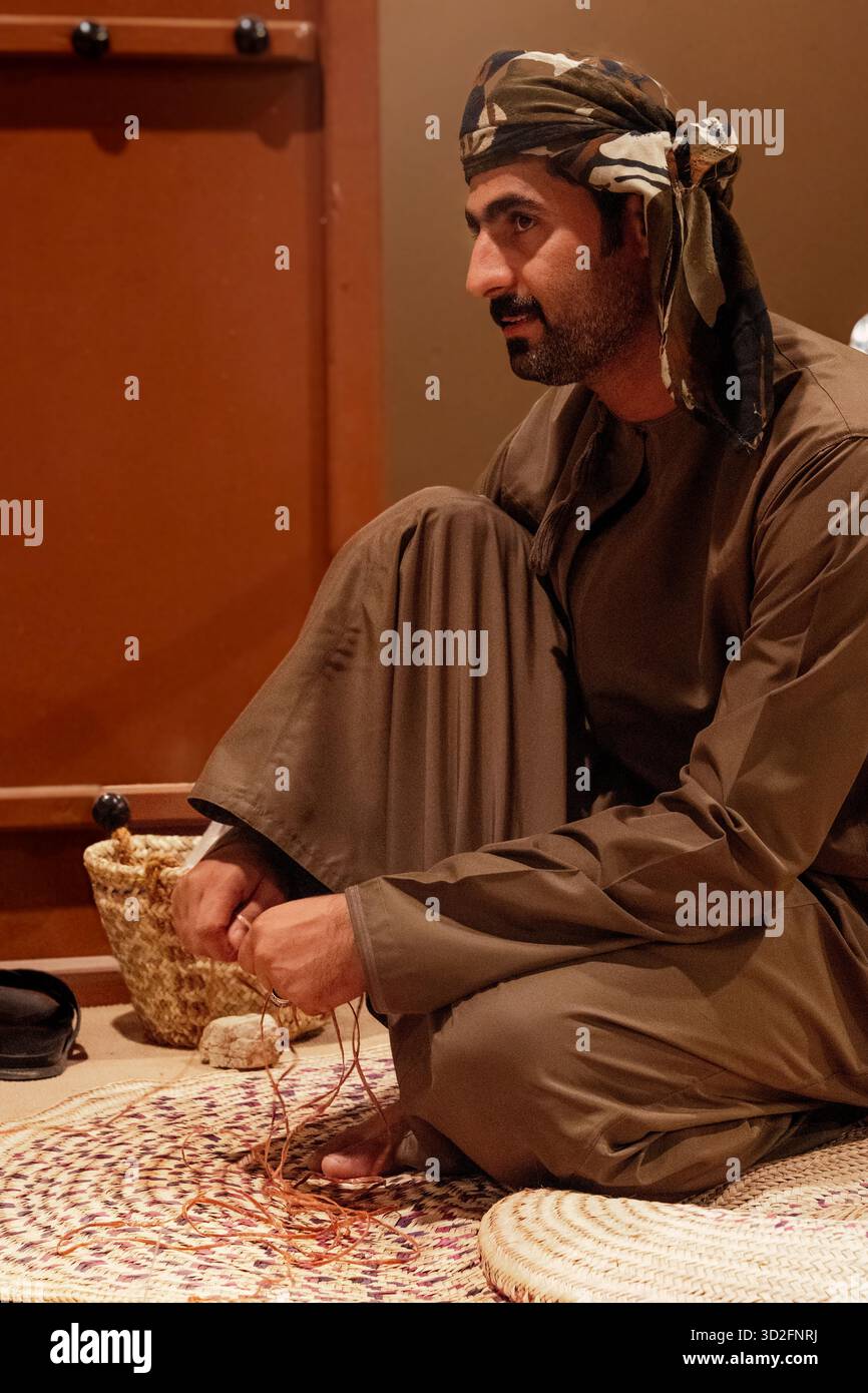 Mascate, Oman - 24 dicembre 2024: Un uomo in tradizionale abbigliamento mediorientale intreccia un cesto o un tappetino utilizzando fibre naturali al Mascate Festival, Oman. Foto Stock