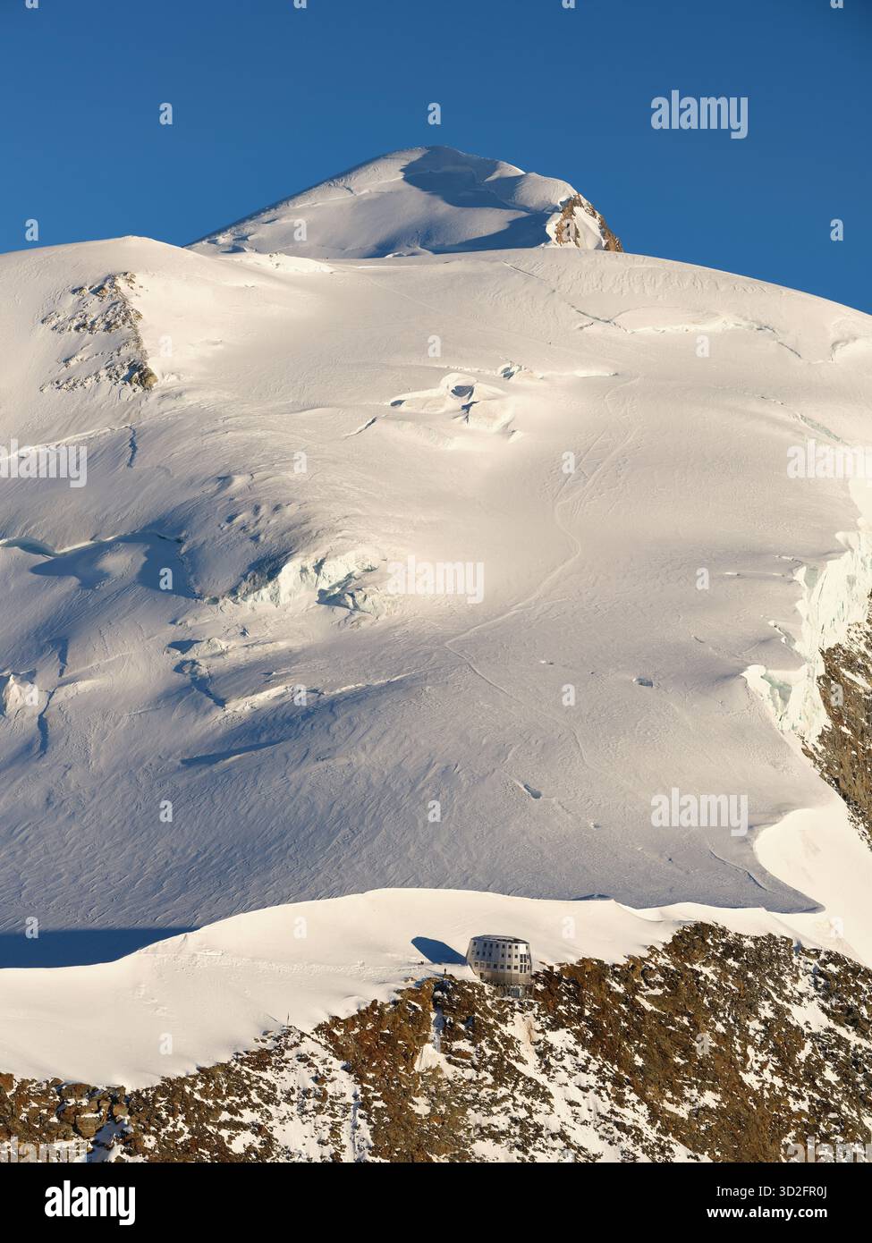 VISTA AEREA. Rifugio Goûter (rifugio più alto della Francia) sulla cima dell'Aiguille du Goûter (3863 m). In lontananza, il Monte bianco (4810 m). Francia. Foto Stock