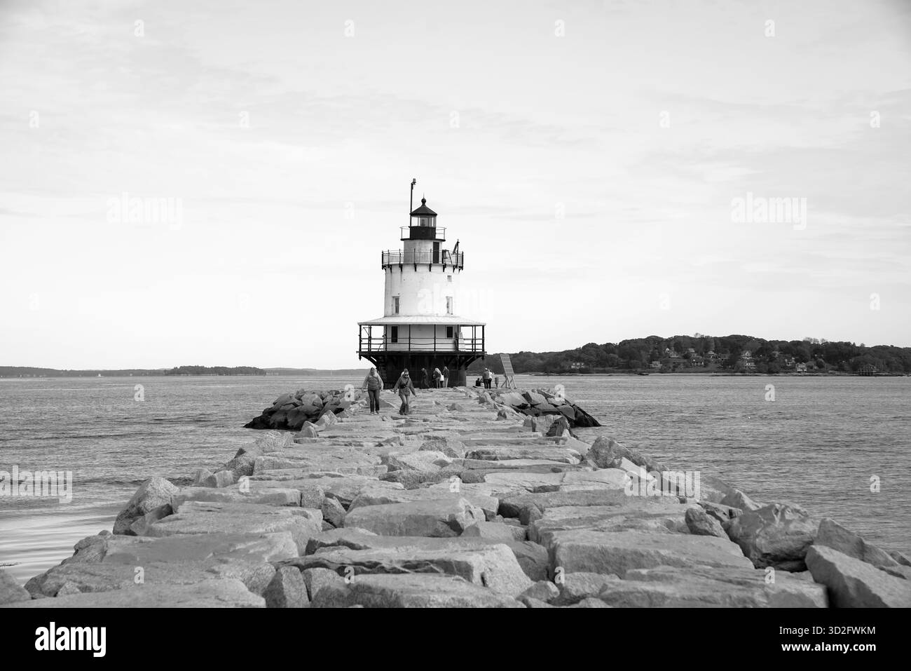 Vista in bianco e nero del faro Spring Point Ledge, con i visitatori che camminano sul molo di pietra sotto cieli sovrastati attraverso casco Bay Foto Stock