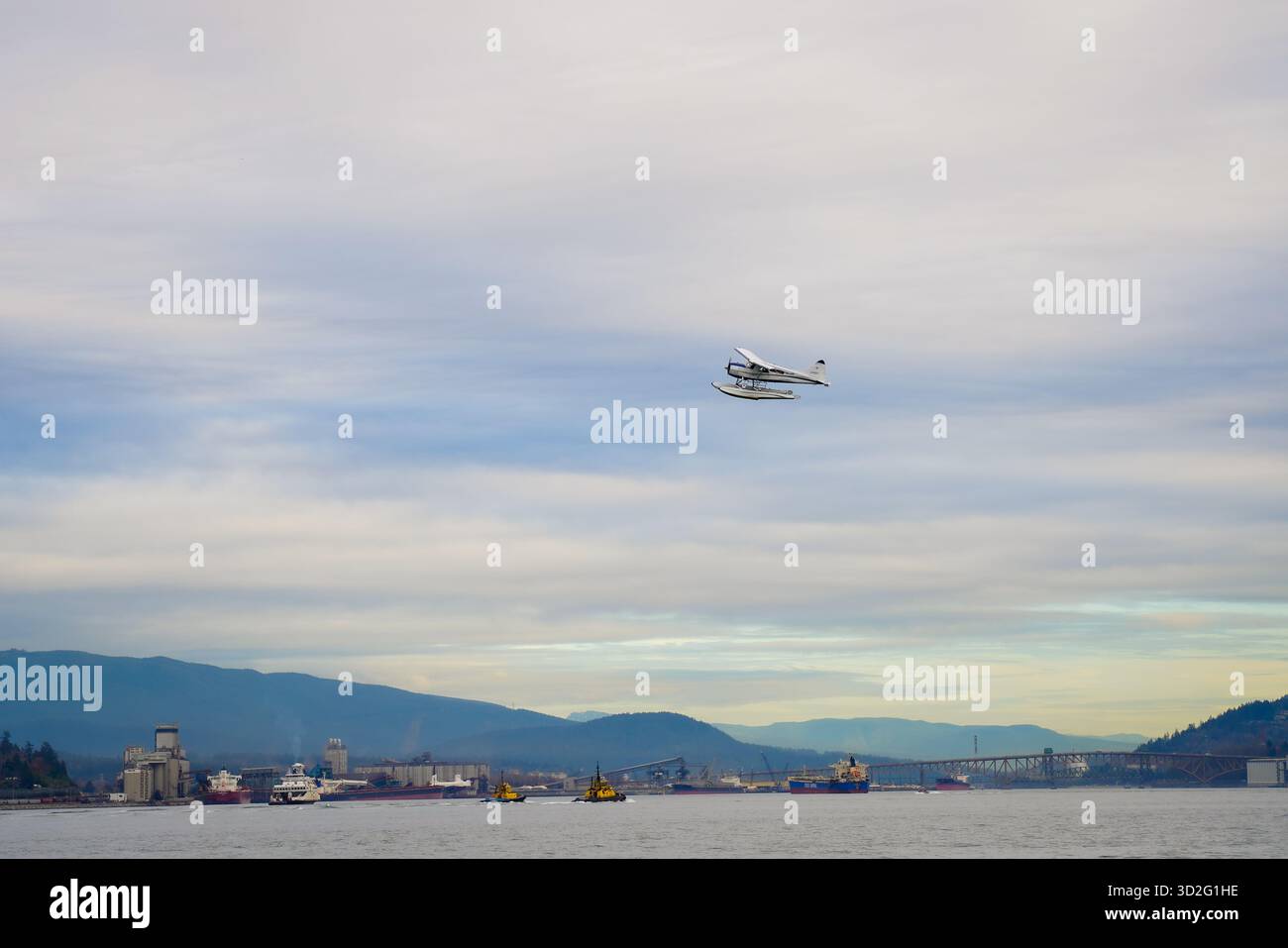Un idrovolante vola sopra Burrard Inlet con navi portuali, terminali industriali e montagne boscose lontane sotto un cielo tenue. Foto Stock