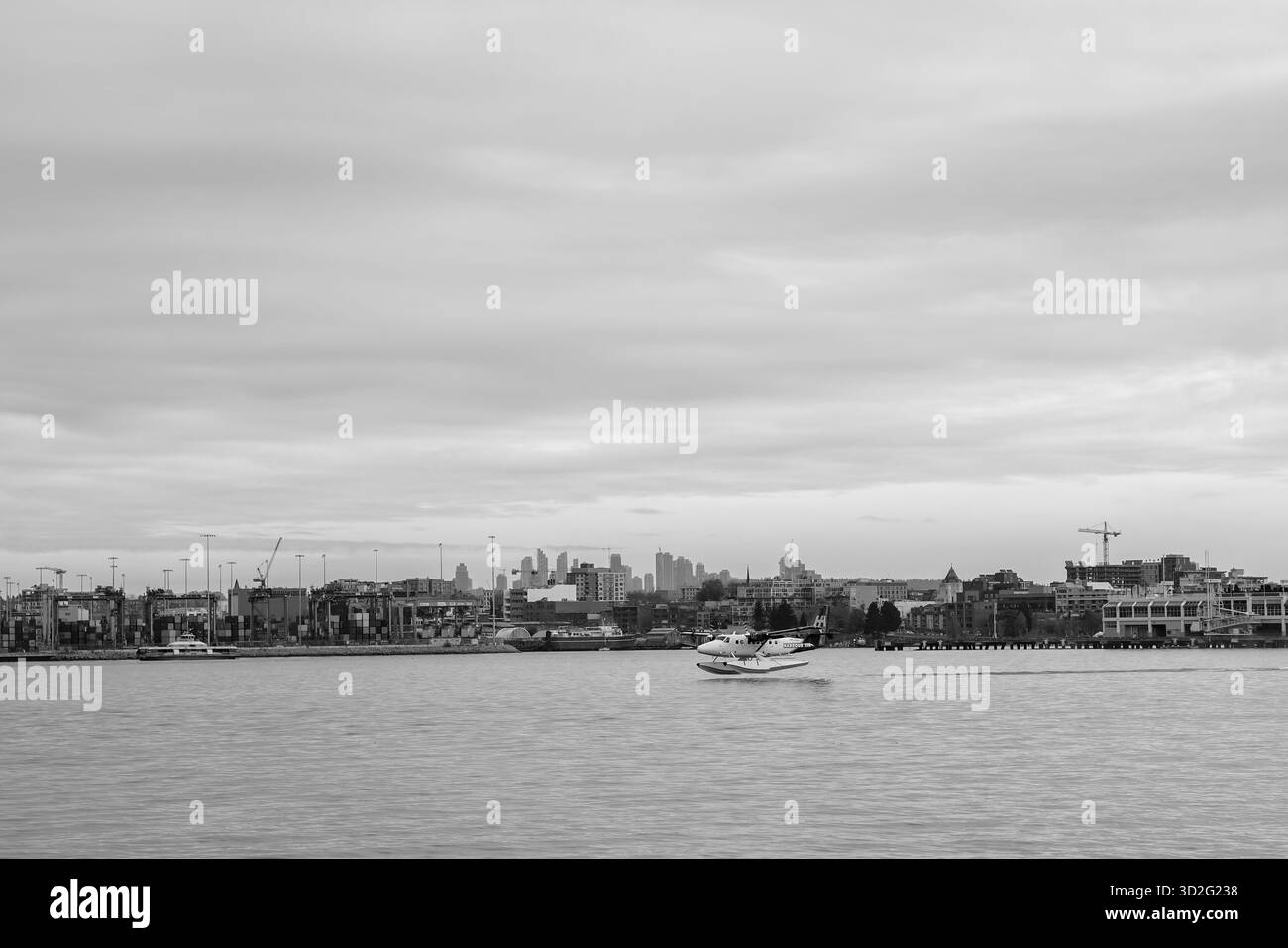 Un taxi in idrovolante Harbour Air lungo il lungomare industriale, con una vista in bianco e nero dei terminal merci e del lontano skyline. Foto Stock