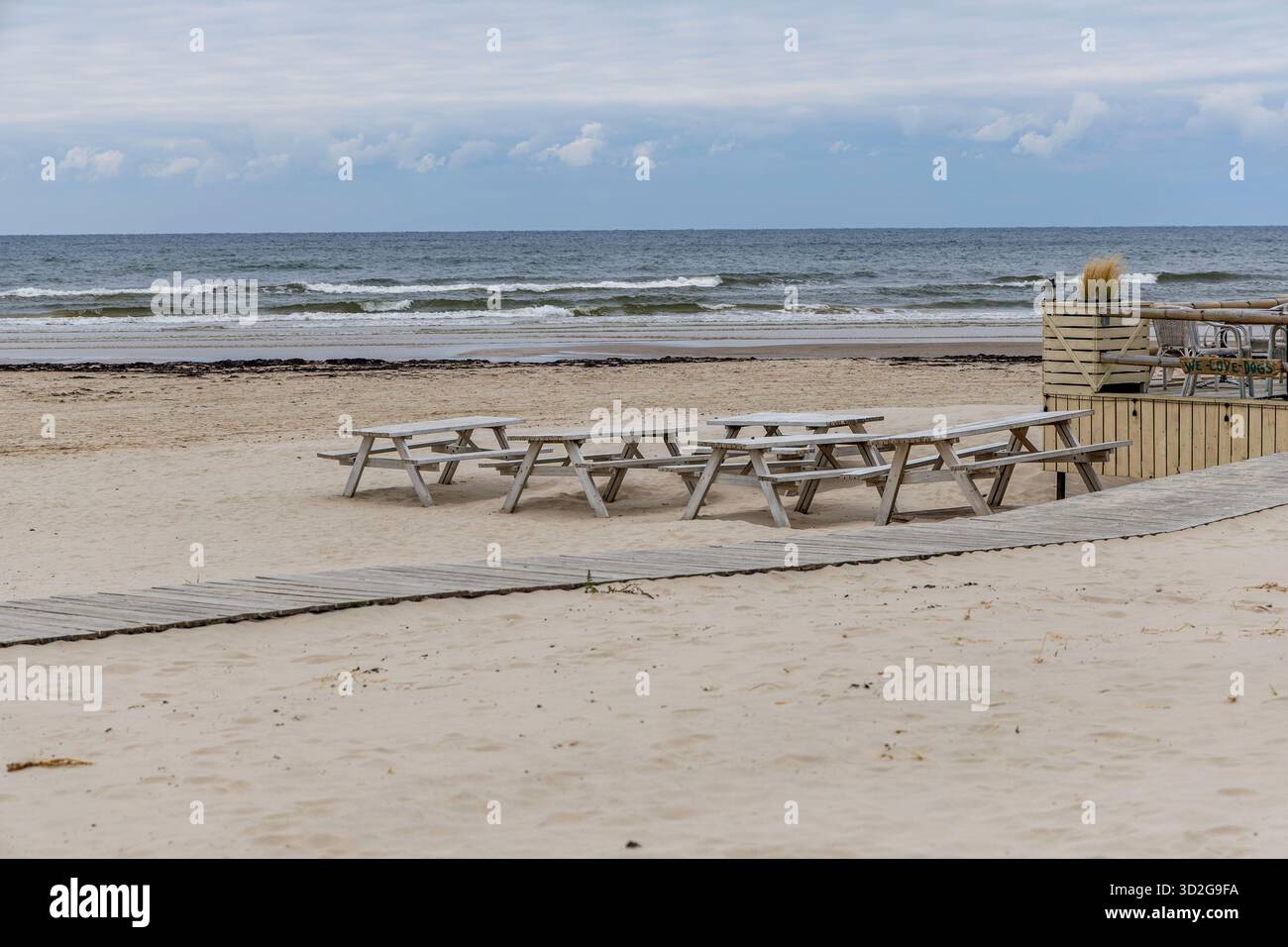 Diverse file di tavoli da picnic in legno seduti su un'ampia e vuota spiaggia di sabbia, con una passerella di legno che conduce ad una terrazza caffè vicino al mare. Foto Stock