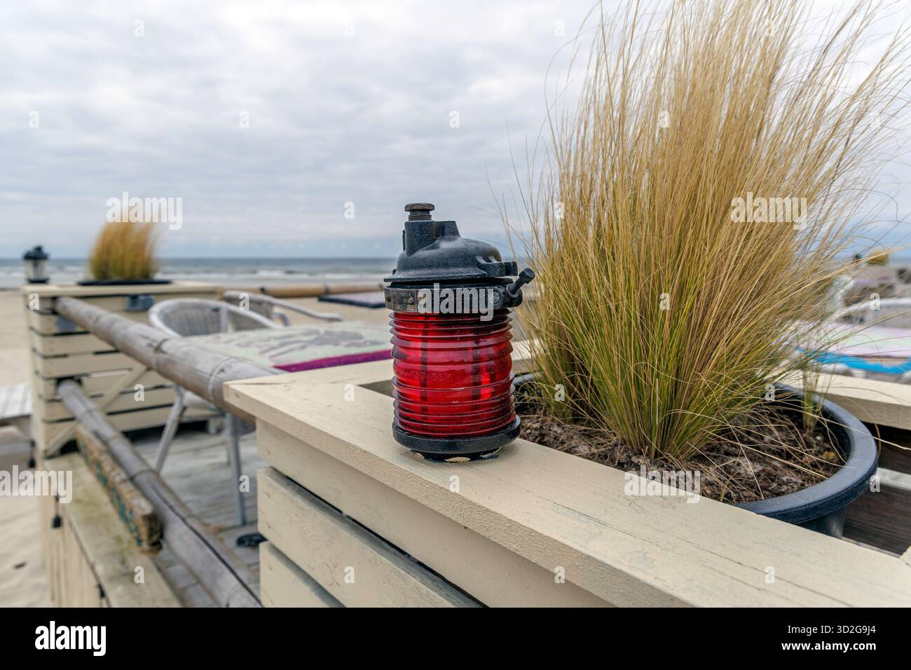 Primo piano di una lanterna marina rossa decorativa accanto a un vaso di alta duna sulla ringhiera di legno di una terrazza vuota sulla spiaggia. Foto Stock