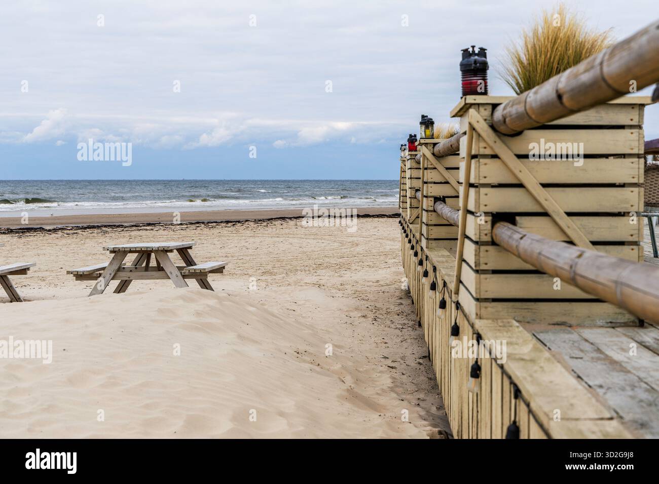 Vista prospettica di una terrazza in legno con lanterne decorative e un tavolo da picnic parzialmente coperto da sabbia soffiata dal vento sulla spiaggia. Foto Stock