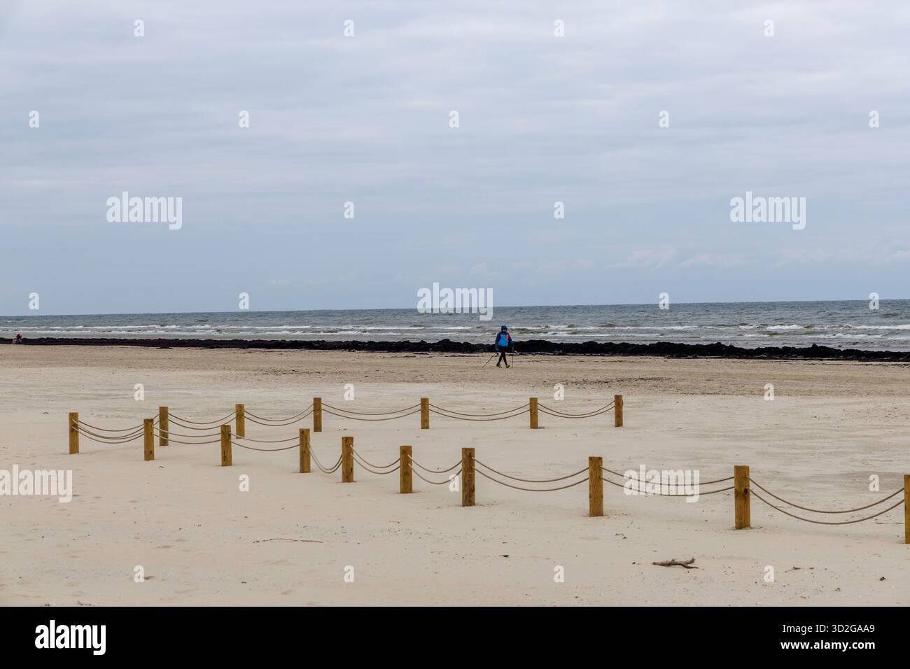Ampia vista panoramica di una vasta spiaggia di sabbia con una barriera decorativa a corda su pali di legno, una persona lontana e una linea di alghe scure vicino al mare sotto a. Foto Stock