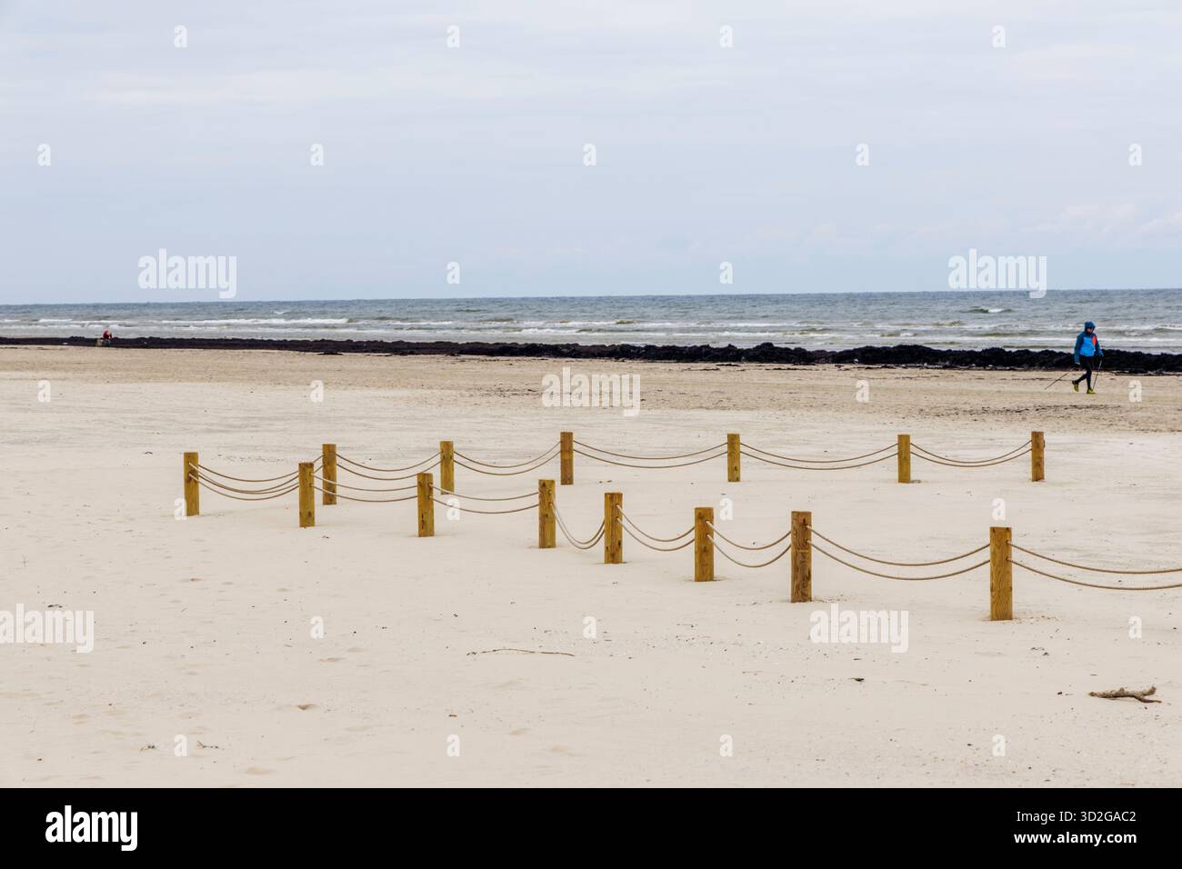 Ampia vista panoramica di una vasta spiaggia di sabbia con una barriera decorativa a corda su pali di legno, una persona lontana e una linea di alghe scure vicino al mare sotto a. Foto Stock