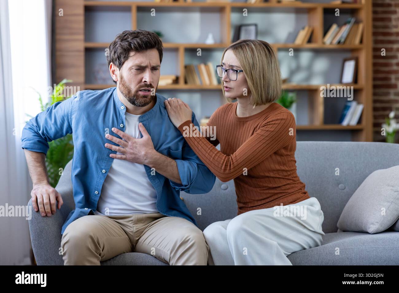 Uomo che soffre di dolore toracico acuto e mancanza di respiro seduto su un divano a casa, ricevendo supporto preoccupato e conforto da una donna durante un'emergenza sanitaria o un attacco di panico Foto Stock