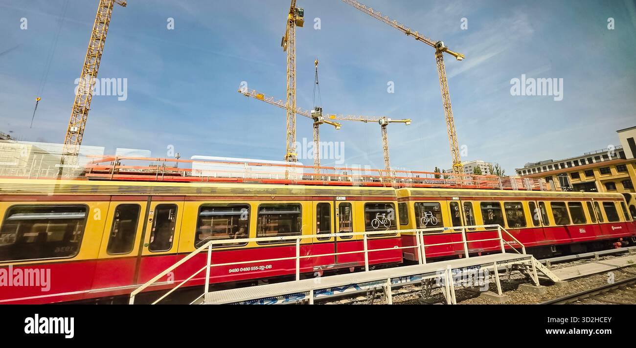 Berlino, Germania - 17 giugno 2025: Il treno rosso e giallo passa accanto a un cantiere con gru che torreggiano sopra. - Immagine stock catturata con smartphone