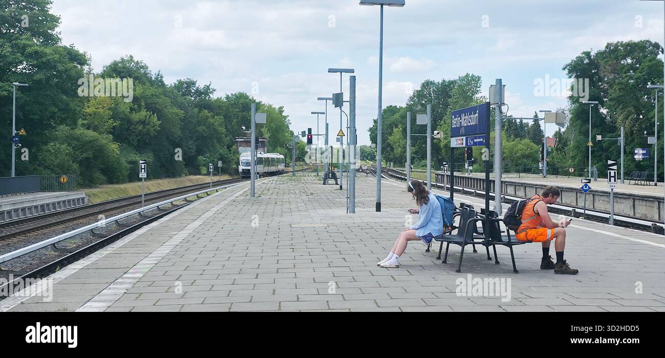 Berlino, Germania - 3 luglio 2025: . Giovani donne e uomini sono seduti sulle panchine della stazione ferroviaria circondati dal verde - Immagine stock catturata con smartphone