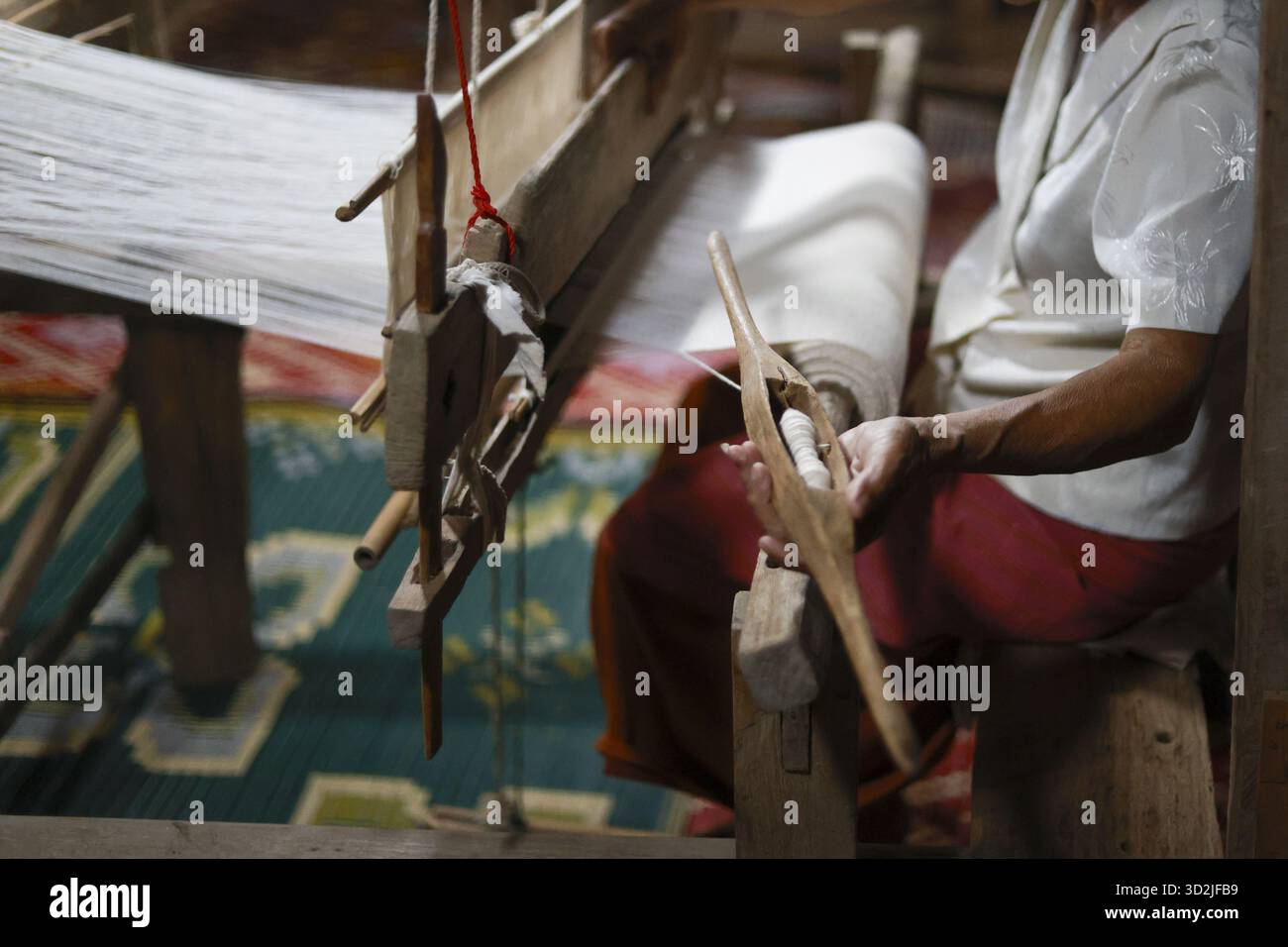 Donna asiatica concentrata che tessera con tradizionale abilità artigianale su telaio in legno. Un artigiano lavora con la navetta e il filo bianco per creare un bellissimo textil Foto Stock