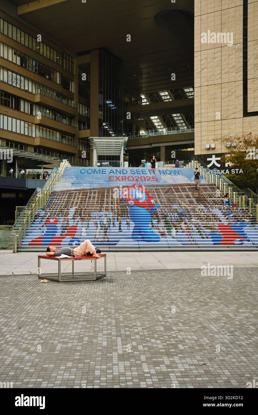Una persona non identificata si rilassa su una panchina di fronte ai gradini decorati della stazione di Osaka, con immagini dell'Osaka Expo e la mascotte Myaku Myaku Foto Stock
