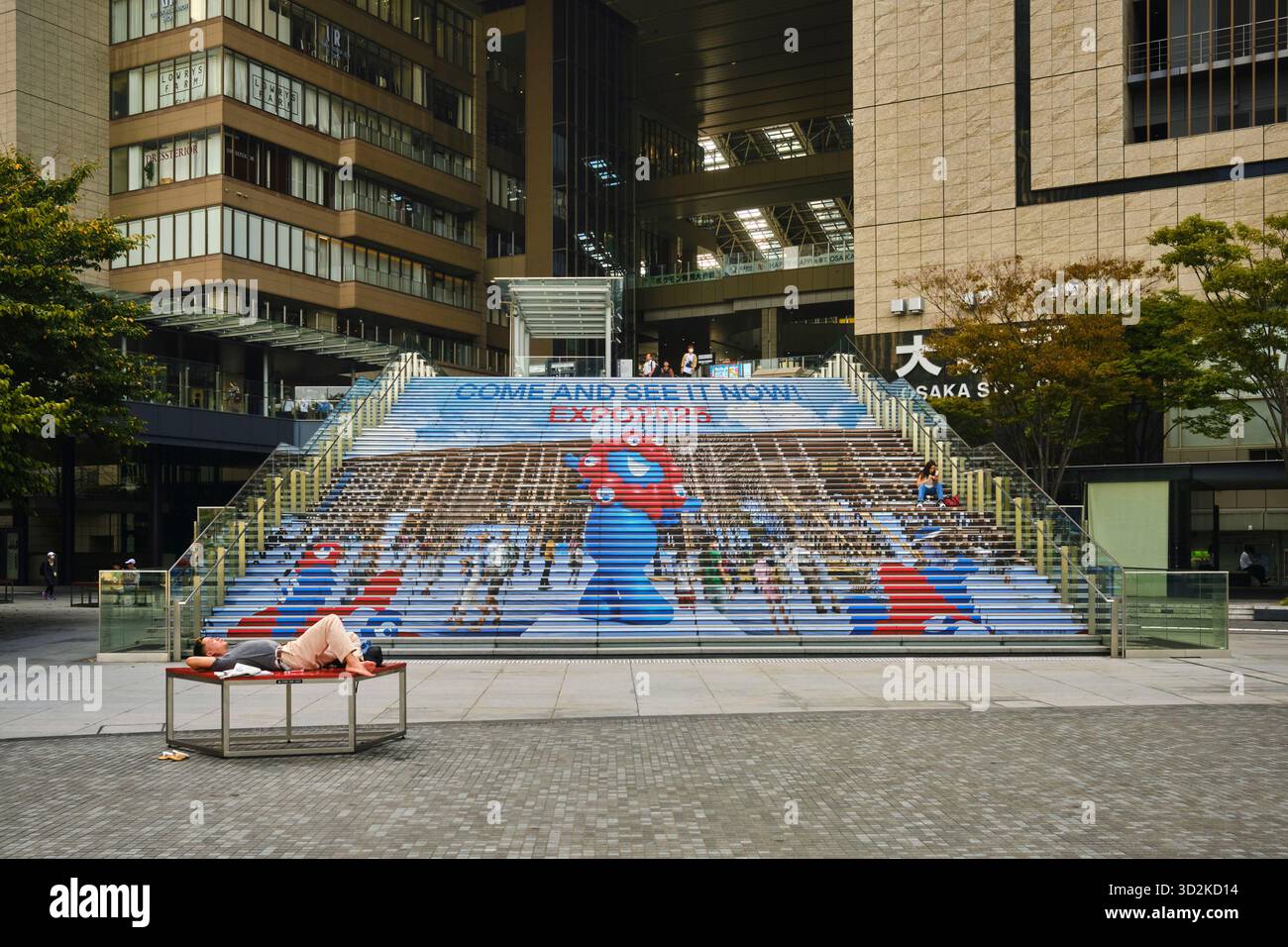 Una persona non identificata si rilassa su una panchina di fronte ai gradini decorati della stazione di Osaka, con immagini dell'Osaka Expo e la mascotte Myaku Myaku Foto Stock