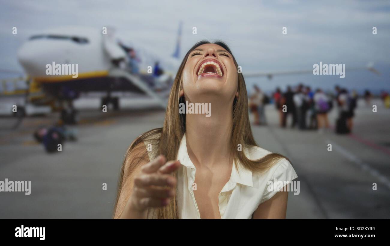 Donna in uniforme da cheerleader che ride con la testa gettata indietro che punta il dito contro l'aereo sull'asfalto dell'aeroporto; gioia. Foto Stock