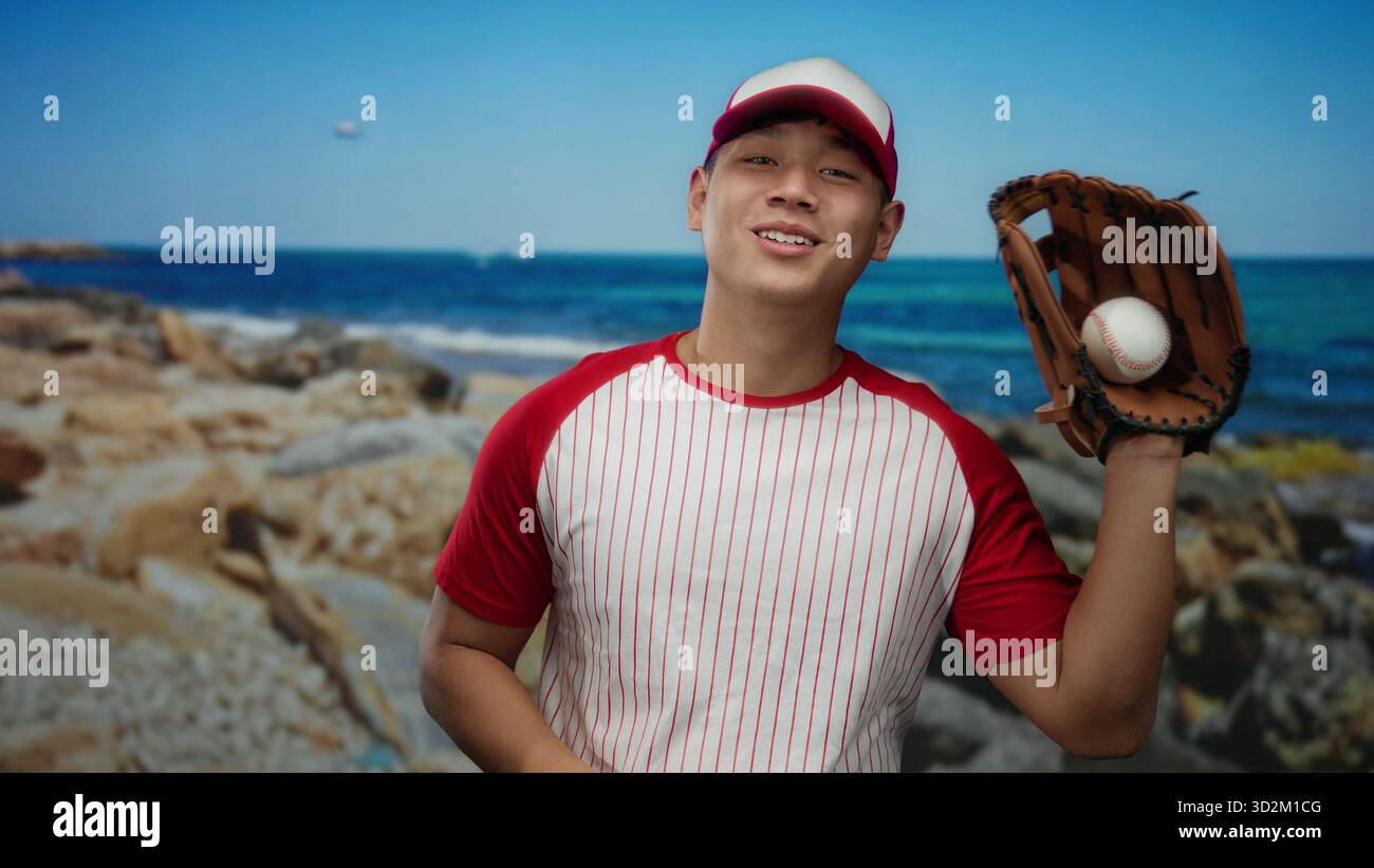 Un giovane con l'uniforme da baseball lancia giocosamente una palla in un pittoresco ambiente sul mare con un oceano cristallino e una spiaggia rocciosa sullo sfondo. Foto Stock