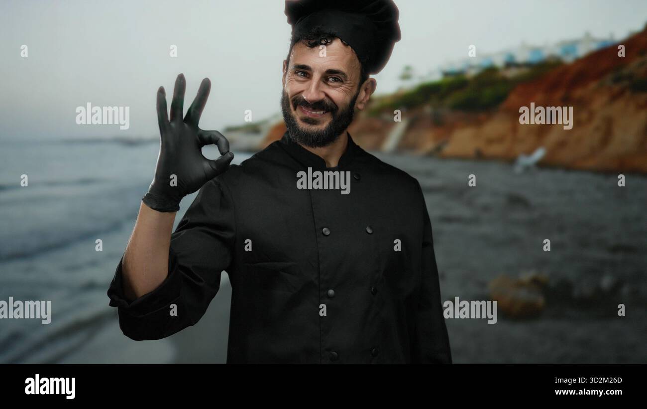 Chef ispanico in uniforme nera che fa un buon gesto con guanti in spiaggia e sullo sfondo del mare, sorridendo suggerendo con sicurezza l'eccellenza culinaria. Foto Stock
